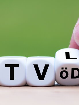 Weiße Würfel mit schwarzen Buchstaben „TVöD“ und „TV-L“ auf Holz, Hand dreht Würfel, grüner Hintergrund