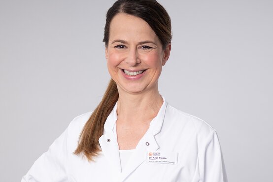 Dr. Katja Staade. Ärztin im weißen Kittel mit Namensschild vor neutral-grauem Hintergrund