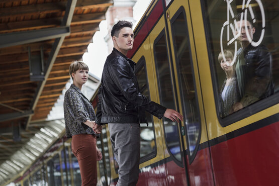 Junge Frau und Mann steigen am Bahnhof in gelb-rote Bahn ein, Spiegelung und Fahrradsymbol im Fenster sichtbar
