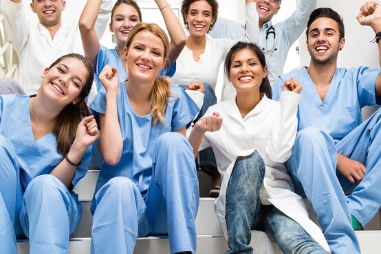 Gruppe junger Menschen in medizinischer Kleidung sitzt lachend und jubelnd auf Treppe in Klinik