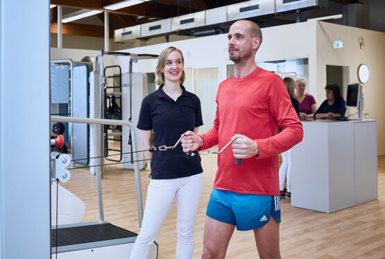 Mann trainiert an Seilzugmaschine, Physiotherapeutin überwacht im hellen Reha- und Gesundheitszentrum