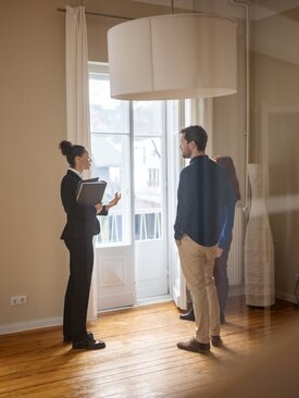 Drei Personen in hellem, leerem Raum mit Holzboden und großen Fenstern, Gesprächssituation