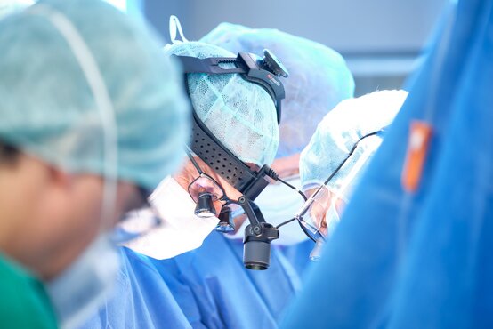 Ärzte in blauen OP-Kitteln und Hauben, Lupenbrille und Stirnlampe, moderne Medizintechnik im Operationssaal