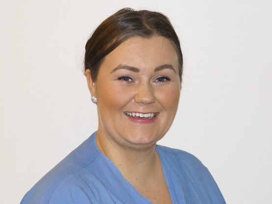 Alina Koy. Freundlich lächelnde Frau im hellblauen Kittel, medizinisches Personal vor weißem Hintergrund