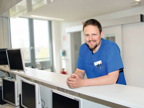 Freundlich lächelnder Mitarbeiter in blauer medizinischer Kleidung an Tresen einer modernen Klinik