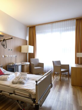 Einzelzimmer in einer Klinik mit Pflegebett, Nachttisch, Sitzbereich, Schreibtisch, Flachbildfernseher, Holzboden und großem Fenster