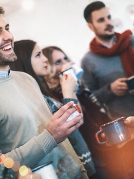 Gruppe junger Erwachsener auf winterlicher Feier, lächelnd im Gespräch, mit Heißgetränken