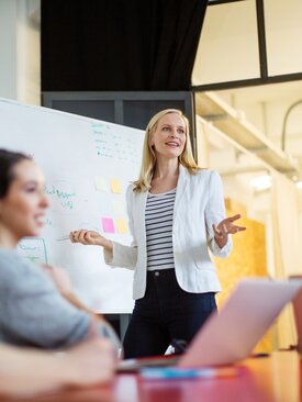 Frau präsentiert vor Whiteboard mit Notizen und Diagrammen im modernen Konferenzraum, drei Personen mit Laptops am Tisch