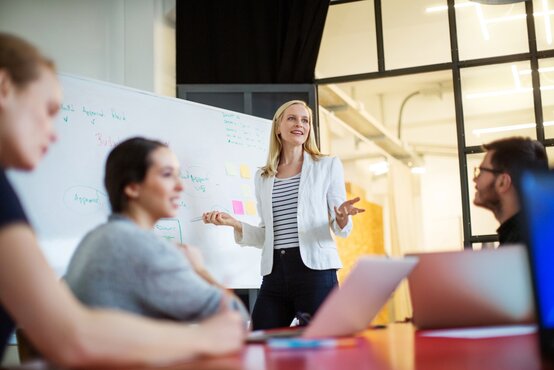 Frau präsentiert vor Whiteboard mit Notizen und Diagrammen im modernen Konferenzraum, drei Personen mit Laptops am Tisch