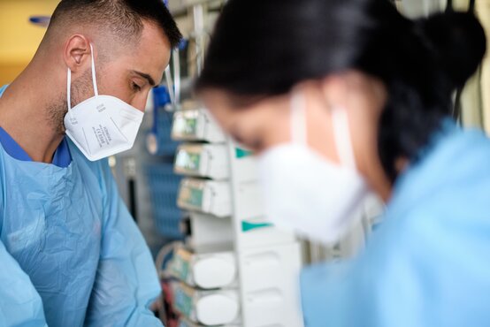 Zwei medizinische Fachkräfte im blauen Kittel und FFP2-Maske arbeiten gemeinsam