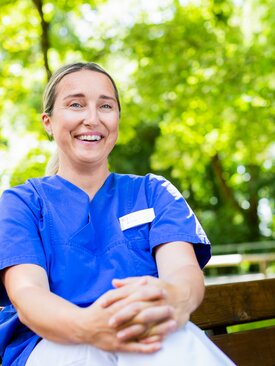 Junge Pflegekraft in blauer Kasack-Uniform sitzt auf einer Bank im grünen