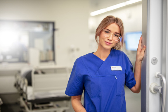Junge Frau in blauer Pflegekleidung mit Namensschild und Brille steht an Tür in hellem Krankenhauszimmer
