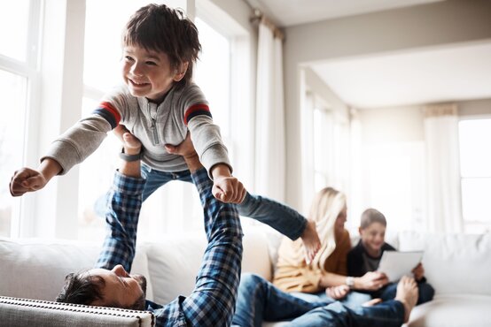 Vater liegt auf einem Sofa und hebt lachendes Kind hoch, im Hintergrund Mutter und weiteres Kind, alle lächeln
