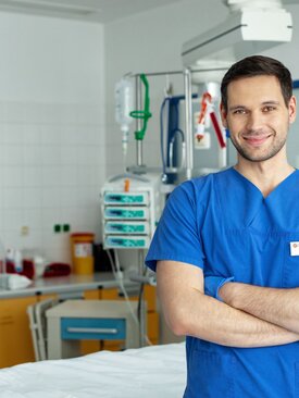 Mann im blauen Pflegekittel steht mit verschränkten Armen in einem modernen Krankenhauszimmer