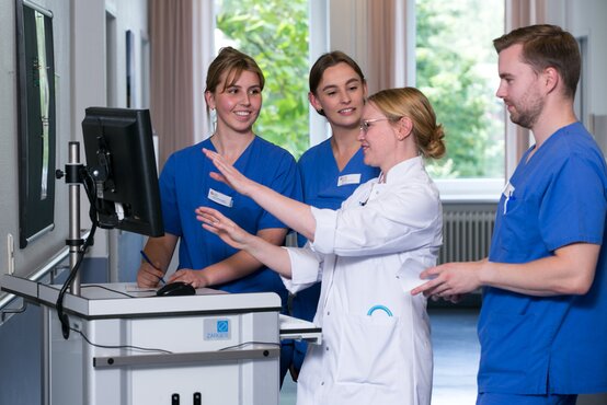 Vier medizinische Fachkräfte stehen in hellem Klinikflur vor Computer, eine Person im weißen Kittel erklärt etwas am Monitor