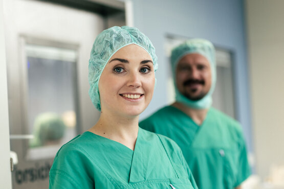Zwei Klinikmitarbeitende in grüner OP-Kleidung und Hauben vor Türen im Krankenhausflur.