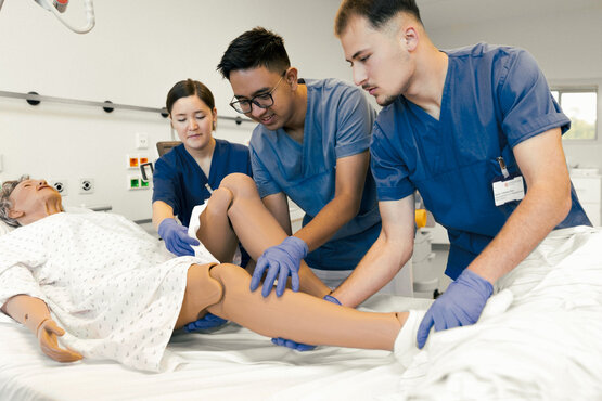 Drei Auszubildende der Schön Klinik Gruppe üben im modernen Schulungsraum mit einer Patientensimulationspuppe im Bett.