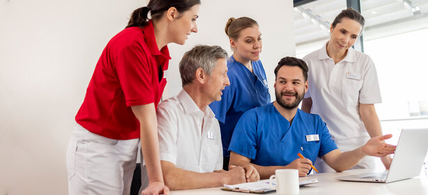 Fünfköpfiges medizinisches Team in Kitteln und rotem Polohemd bespricht sich am Tisch mit Laptop und Notizblock im hellen Raum