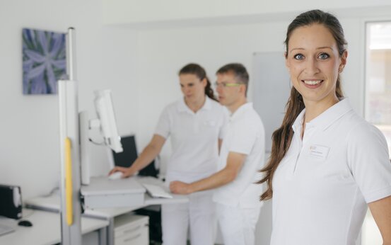 Drei Personen in weißer Kleidung, eine Frau mit Pferdeschwanz im Vordergrund, zwei am Schreibtisch mit Monitor im hellen, modernen Praxisraum