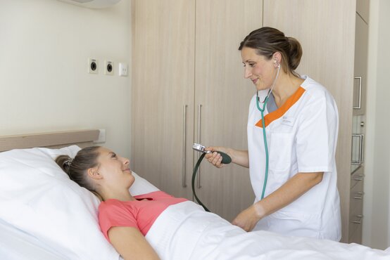 Pflegekraft misst lächelnder Patientin im Bett den Blutdruck mit Messgerät in heller, freundlicher Krankenhausumgebung