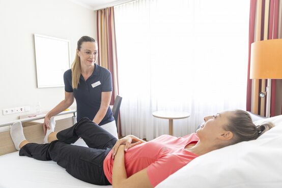 Physiotherapeutin in dunkelblauem Polo-Shirt behandelt das Bein einer Patientin im rosa T-Shirt auf Klinikbett