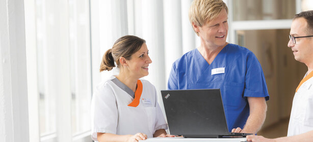 Drei Pflegekräfte im Krankenhausflur besprechen gemeinsam digitale Patientendaten am Laptop, Teamarbeit im Gesundheitswesen