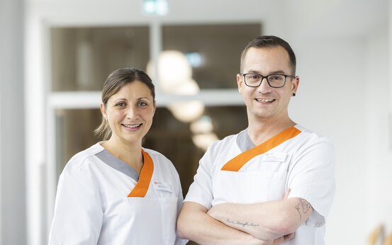 Zwei Pflegekräfte in weißer Dienstkleidung mit orange-grauen Akzenten lächeln im modernen, hellen Klinikbereich