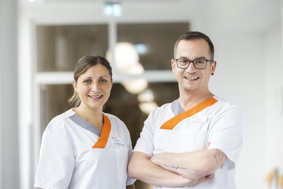 Zwei Pflegekräfte in weißer Dienstkleidung mit orange-grauen Akzenten lächeln im modernen, hellen Klinikbereich