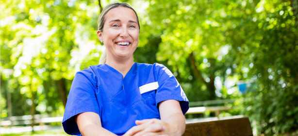 Junge Pflegekraft in blauer Kasack-Uniform sitzt auf einer Bank im grünen