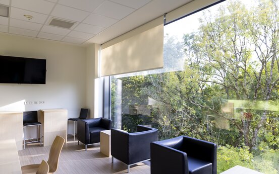 Heller Aufenthaltsraum mit bodentiefen Fenstern, schwarzen Sesseln, Holztischen und Blick ins Grüne