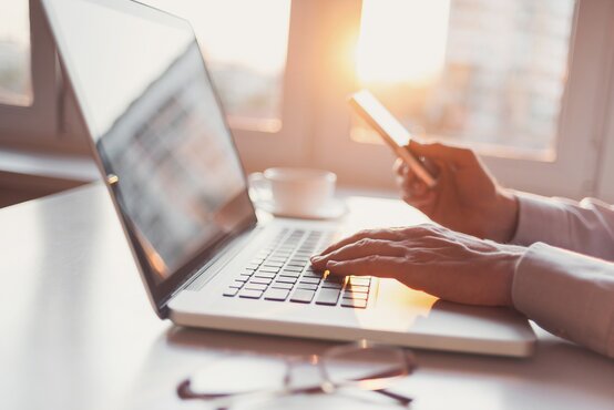 Mann im hellen Hemd arbeitet am Laptop und hält Smartphone, Kaffeetasse und Brille auf hellem Schreibtisch, Fenster im Hintergrund