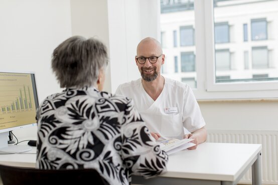 Arzt im weißen Kittel spricht am Tisch mit älterem Patienten in heller Klinik