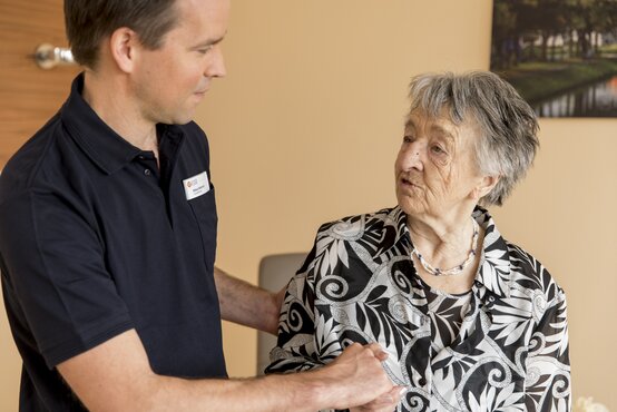 Physiotherapeut hält Hand einer älteren Frau mit grauen Haaren im hellen Raum, beide stehen sich zugewandt