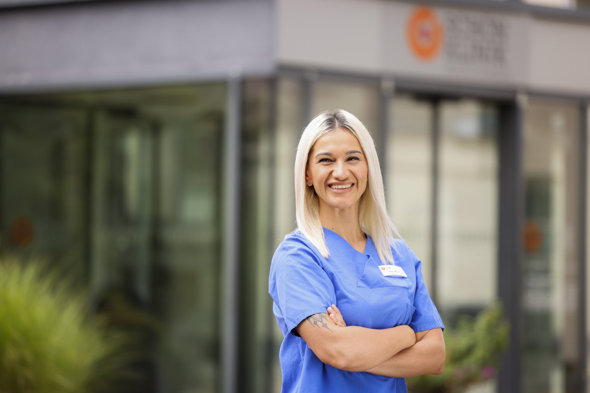 Junge Frau mit blonden Haaren in blauer Pflegekleidung und Namensschild steht lächelnd vor Schön Klinik München Schwabing