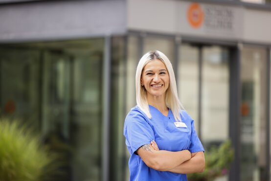 Junge Frau mit blonden Haaren in blauer Pflegekleidung und Namensschild steht lächelnd vor Schön Klinik München Schwabing