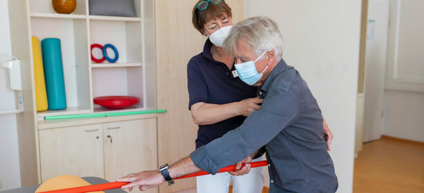 Mann mit Maske hält roten Gymnastikstab, Therapeutin mit Maske unterstützt im hellen Therapieraum