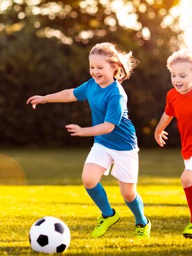Zwei fröhliche Kinder spielen gemeinsam Fußball auf grünem Sportplatz bei Sonnenschein