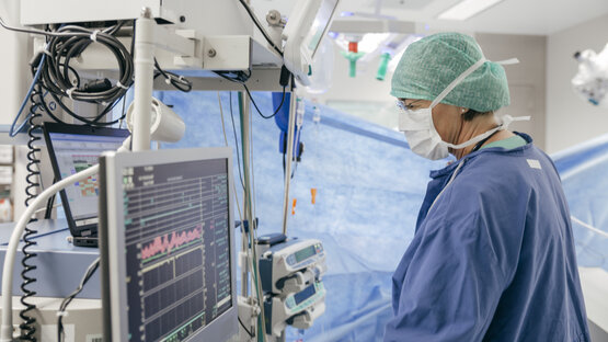 Ärztin in blauer OP-Kleidung und Mundschutz bedient Narkosegerät im Operationssaal, Monitor und Beatmungsgerät im Hintergrund