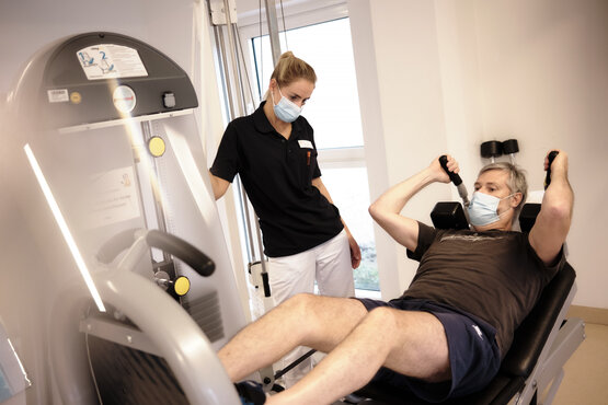 Physiotherapeutin hilft Patientin beim Training an Trainingsgerät in hellem Therapieraum mit großen Fenstern