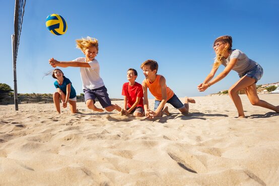 Fünf Kinder spielen am sonnigen Sandstrand Volleyball, springen und knien im Sand, Volleyballnetz und Dünen im Hintergrund