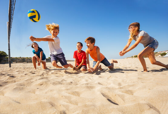 Fünf Kinder spielen am sonnigen Sandstrand Volleyball, springen und knien im Sand, Volleyballnetz und Dünen im Hintergrund