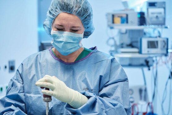 Medizinisches Fachpersonal in blauer Schutzkleidung, Haube und Mund-Nasen-Schutz hält Instrument im OP, medizinische Geräte im Hintergrund