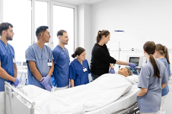 Gruppe von Pflegeauszubildenden in blauer Kleidung steht im modernen Patientenzimmer um ein Krankenbett mit Simulationspuppe, Ausbilderin erklärt medizinischen Monitor, praxisnahes Lernen und Teamarbeit in der Pflegeausbildung