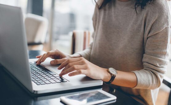 Junge Frau schreibt konzentriert am Laptop eine Bewerbung für den Pflegeberuf, Smartphone auf hellem, ordentlichem Tisch