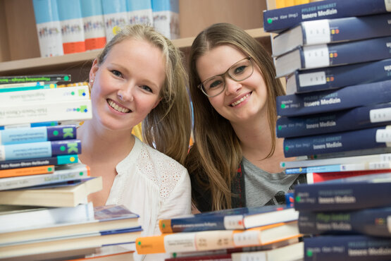 Zwei junge Frauen zwischen Bücherstapeln, lächelnd, mit Büchern wie „Pflege Kompakt Innere Medizin“.