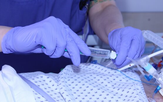 Pflegekraft in blauer Kleidung und Handschuhen führt mit Spritze und Beatmungsschlauch eine Intensivpflege-Maßnahme durch