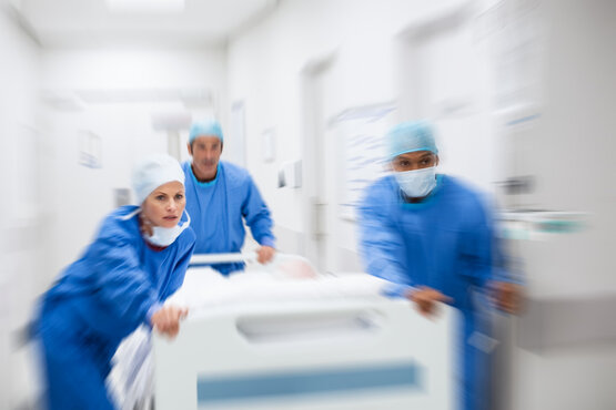 Drei Pflegekräfte in blauer Schutzkleidung schieben eine Patiententrage eilig durch einen Krankenhausflur.
