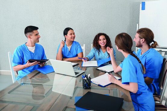 Fünf junge Erwachsene in medizinischer Kleidung sitzen an einem Tisch, lachen und lernen gemeinsam, Bücher und Notizblöcke vor sich