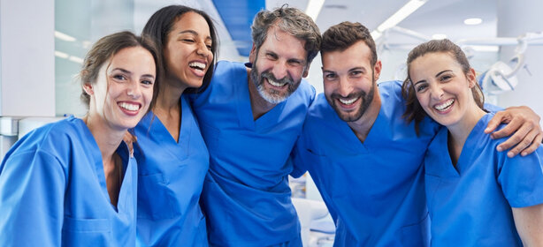 Fünf Personen in blauen Kasacks stehen eng zusammen, lächeln und befinden sich in einer hellen Klinik