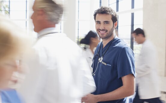 Mann im blauen Kittel mit Stethoskop lächelt, weitere Personen in Krankenhauskleidung im Hintergrund